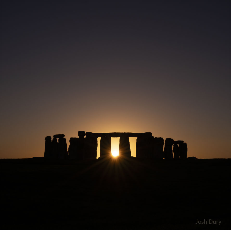 The sun is setting across a field in a clear sky.
In the field are the famous stones of Stonehenge. The
Sun peaks out from the center of the stone array.
Please see the explanation for more detailed information.