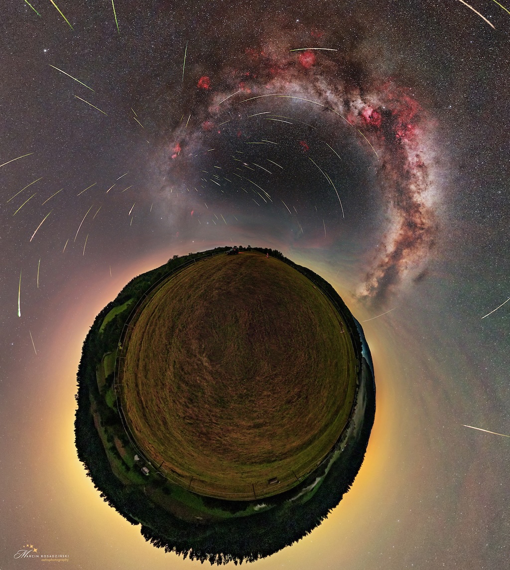 The ground is shown in a little planet perspective, surrounded by the sky with a diffuse glow. The Milky Way is visible in the sky, as well as some nebulae in red and over a hundred meteors. Most of them appear to come from the same point.