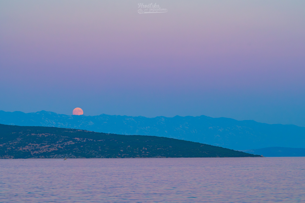 Über dem Meer ragen Berge der Insel Krk auf, über denen der Vollmond aufgeht. Zusammen mit dem Mond steigt auch ein bleigraues Band auf, das sich nach oben hin rosarot verfärbt. Es ist der Schatten der Erde, der oben in den Venusgürtel übergeht.