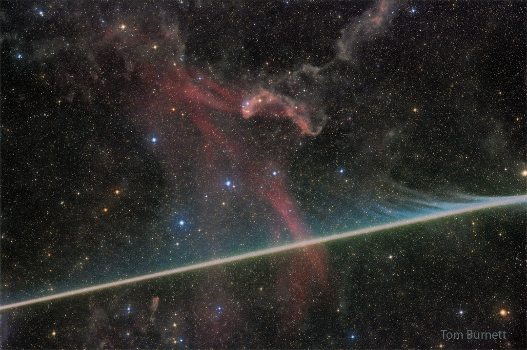 A dark starfield has a faint red nebula near 
the center. A bright streak crosses the image 
horizontally from the lower left, filled with faint
evaporating wisps in many colors. 
Please see the explanation for more detailed information.