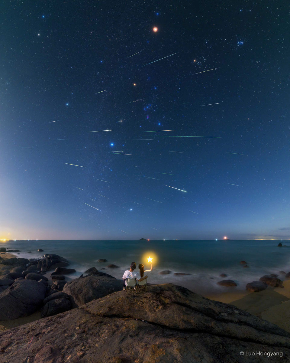 The featured image is a composite showing many meteors trails
streaking across a sky featuring the familiar constellation of Orion.
In the foreground two people sit in adjoining chairs facing away
from the camera, one holding a wand with a glowing star at the end.
Please see the explanation for more detailed information.