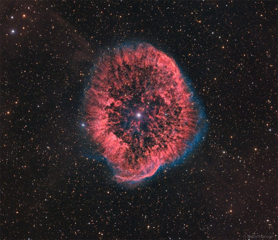A red oval and textured nebula is seen surrounded by a faint
blue glow. A bright star is visible in the center, and many faint
stars are visible in the background. 
Please see the explanation for more detailed information.