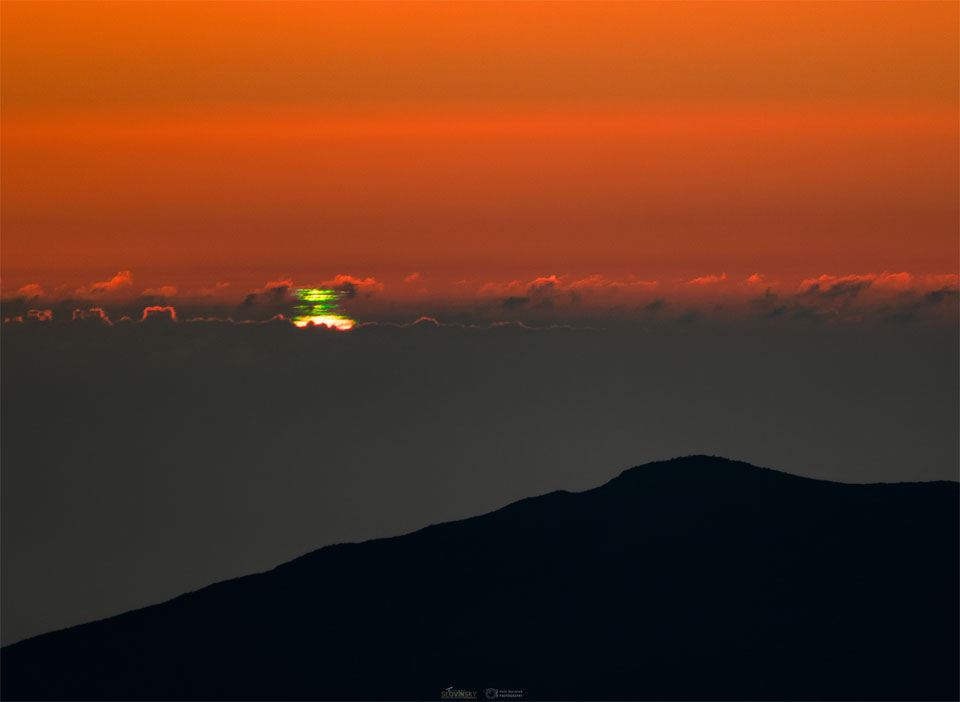 A distant sunset is seen between an orange sky and
dark clouds. A close look at the Sun shows it is topped 
with several green strips, each known as a green flash.
Please see the explanation for more detailed information.