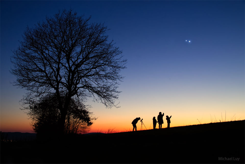 Two bright objects appear in a sky over a hill. 
On the hill, the silhouettes of several people are visible,
including a person looking though a telescope and what
appears to be two children looking upward.
Please see the explanation for more detailed information.