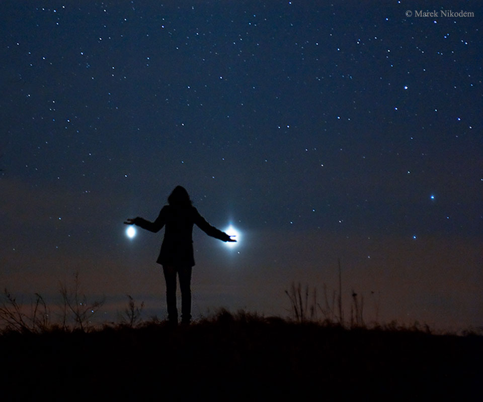 Two bright spots are seen on either side of a person
standing on a hill who appears to be holding one or both of them. 
A starry sky appears in the background.
Please see the explanation for more detailed information.