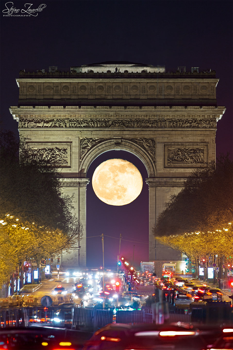 A nearly full Moon is seen through the famous Arc de Triomphi
with trees and cars lining the foreground.
Please see the explanation for more detailed information.