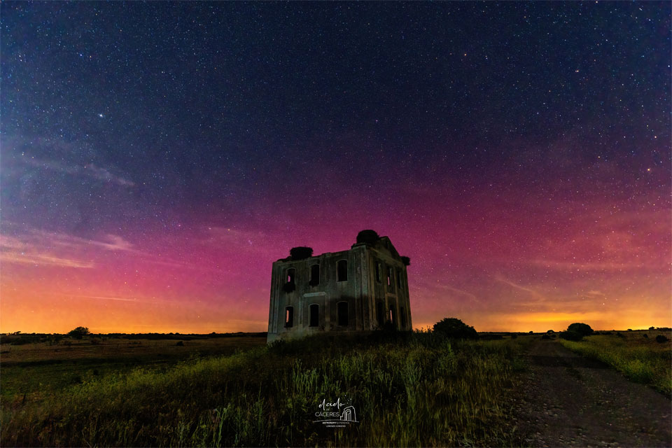 A dark rectangular building is seen across a green field  with colorful aurora, mostly red, seen in the background sky.   Past the aurora, the sky is also filled with stars.   Please see the explanation for more detailed information.