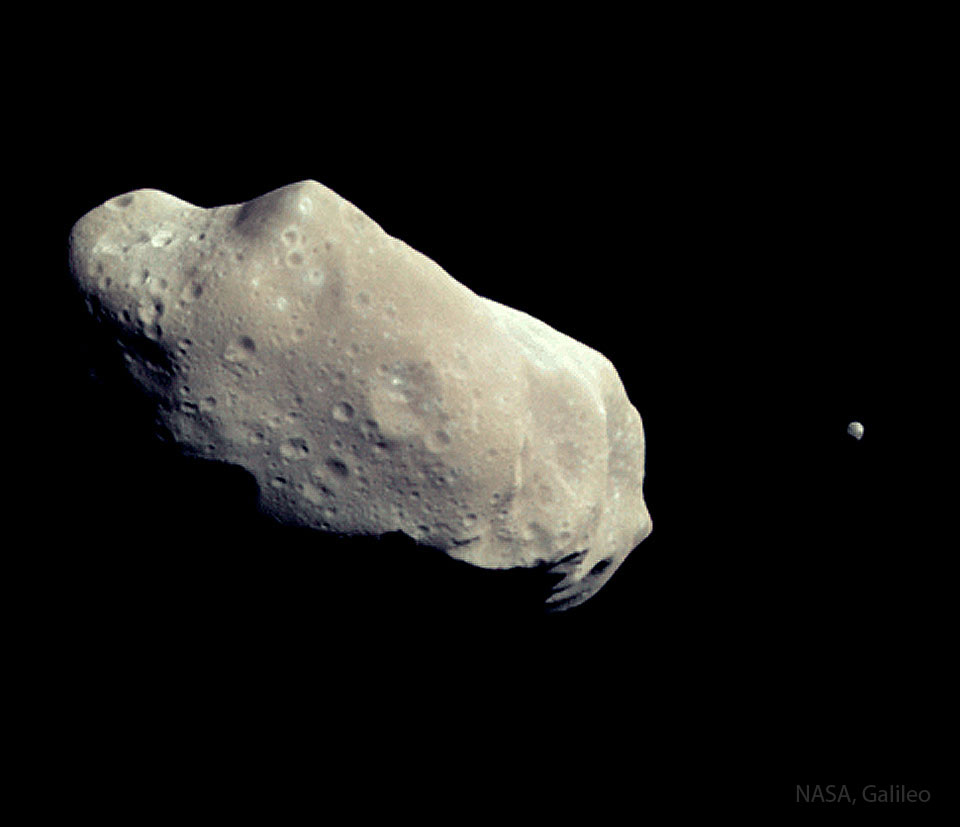 A pair of asteroids are shown with a large, elongated and cratered
one on the left and a much smaller one on the far right.
Please see the explanation for more detailed information.