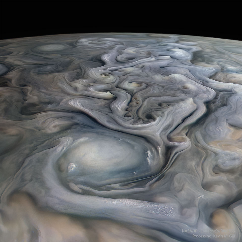 The cloud tops of Jupiter are pictured in a closeup flyby of the Juno spacecraft. A big white oval cloud is visible in the foreground, while many swirls of many muted colors are visible trailing behind. A dark night sky is in the background. Please see the explanation for more detailed information.