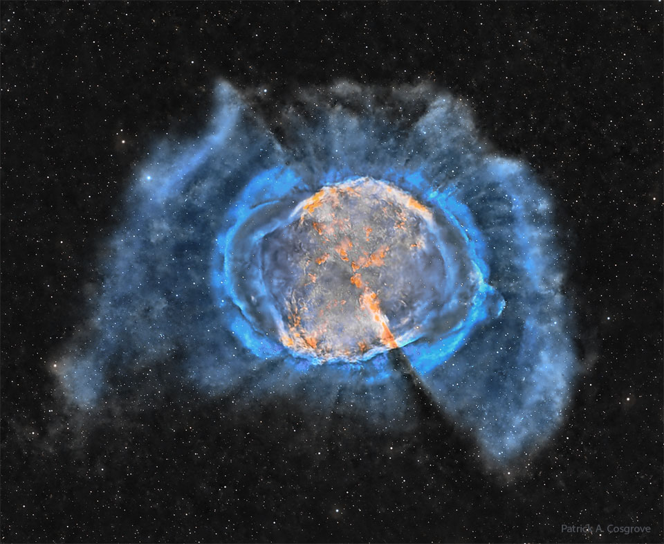 An expansive interstellar gas cloud is shown with an orange interior and outer blue filaments. Many stars are visible in the dark background. Please see the explanation for more detailed information.