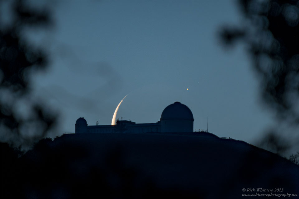 A dark mountain lies in the center with an observatory
building sporting two telescope domes. The background sky
appears dark blue. Behind the center of the observatory
is part of a crescent moon, with an unusual bright spot
to its upper left.
Please see the explanation for more detailed information.