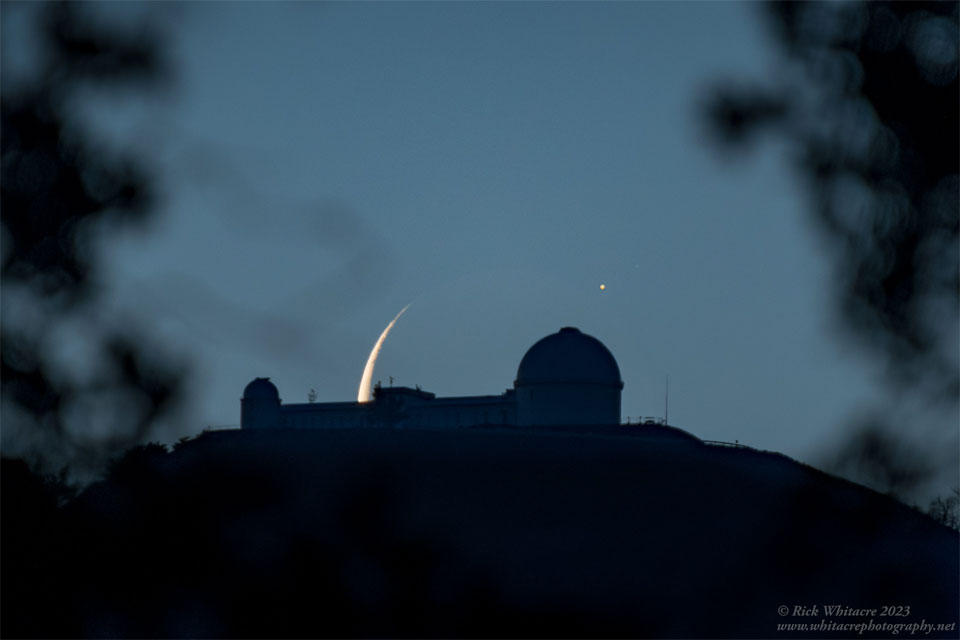 A dark mountain lies in the center with an observatory  building sporting two telescope domes. The background sky  appears dark blue. Behind the center of the observatory  is part of a crescent moon, with an unusual bright spot  to its upper left.  Please see the explanation for more detailed information.