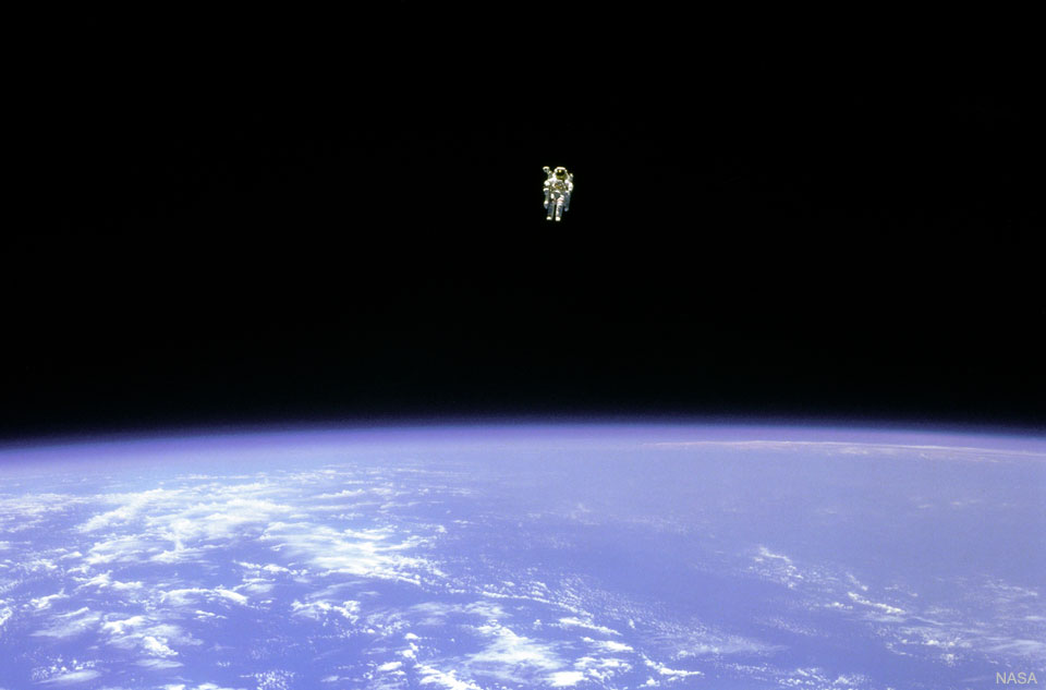 An astronaut is seen hovering over the Earth. In the   top part of the image, the astronaut is seen against the   darkness of space. In the lower part of the image, the  Earth is bright blue with white clouds.    Please see the explanation for more detailed information.