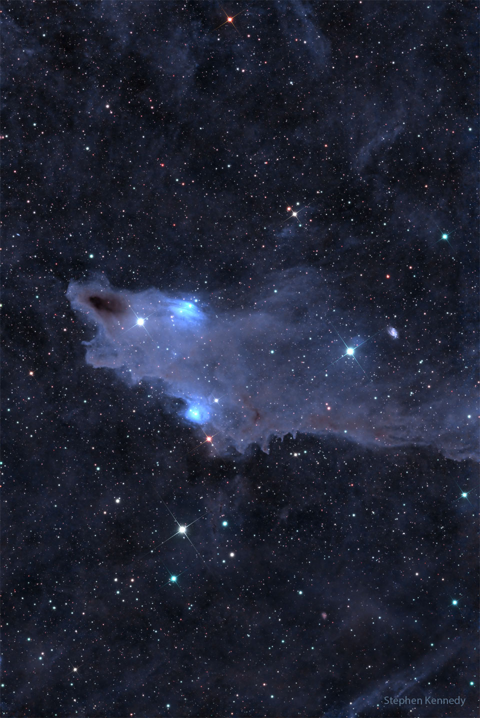 A dark brown cloud that appears similar to a shark is seen
against a background filled with stars and less prominent 
blue-shaded nebulas.
Please see the explanation for more detailed information.