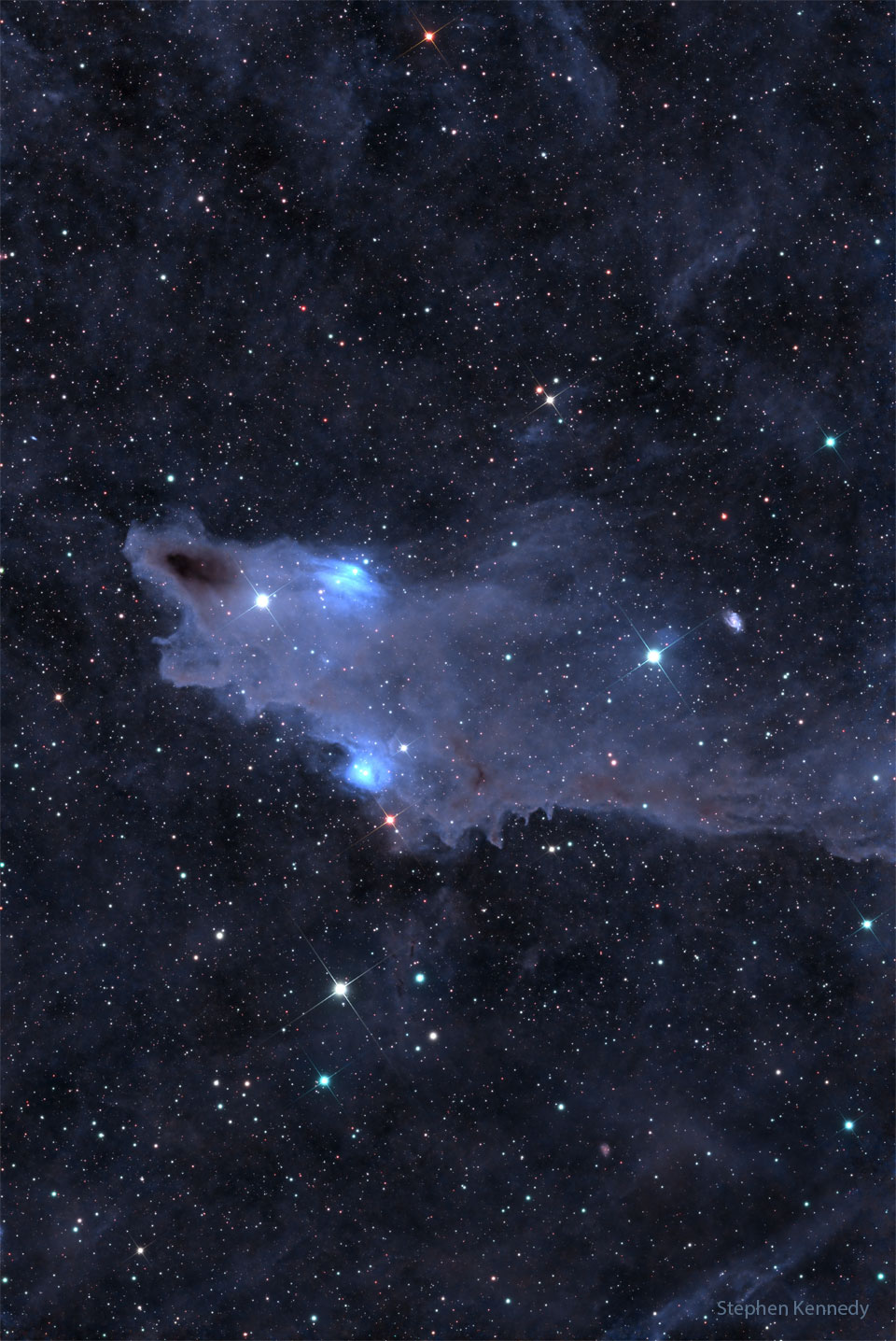 A dark brown cloud that appears similar to a shark is seen  against a background filled with stars and less prominent   blue-shaded nebulas.  Please see the explanation for more detailed information.