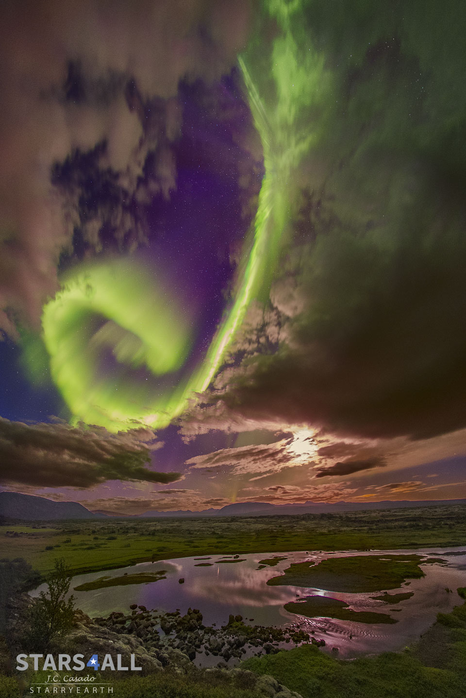A bright green spiral aurora is seen in a break in the 
clouds before a purple background. The foreground contains 
green grassland and a circular lake.
Please see the explanation for more detailed information.