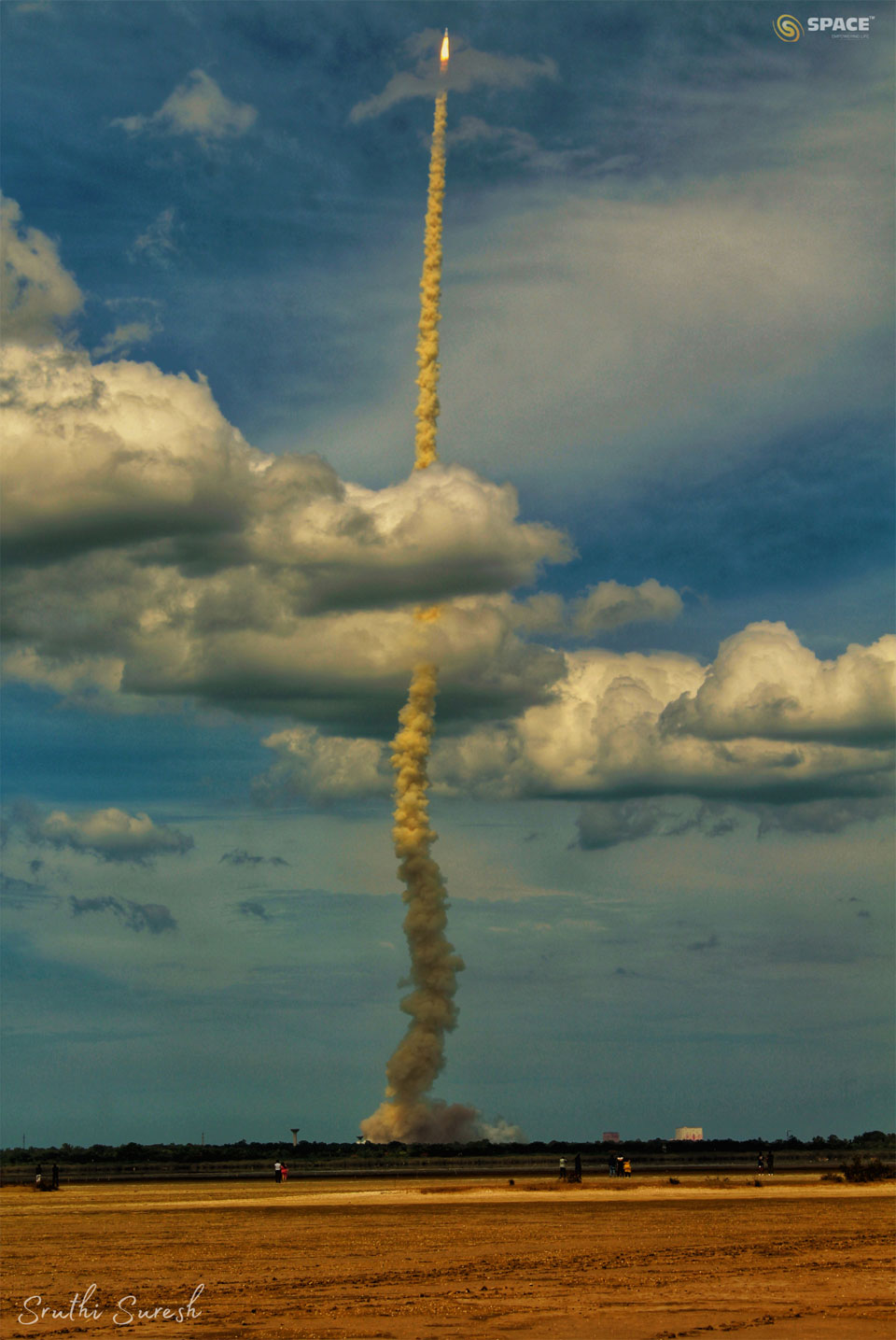 A rocket is seen after lift-off with a long smoke plume.
The rocket is captured against a blue sky and has gone through
a cloud deck. In the foreground is an empty tan-colored field. 
Please see the explanation for more detailed information.