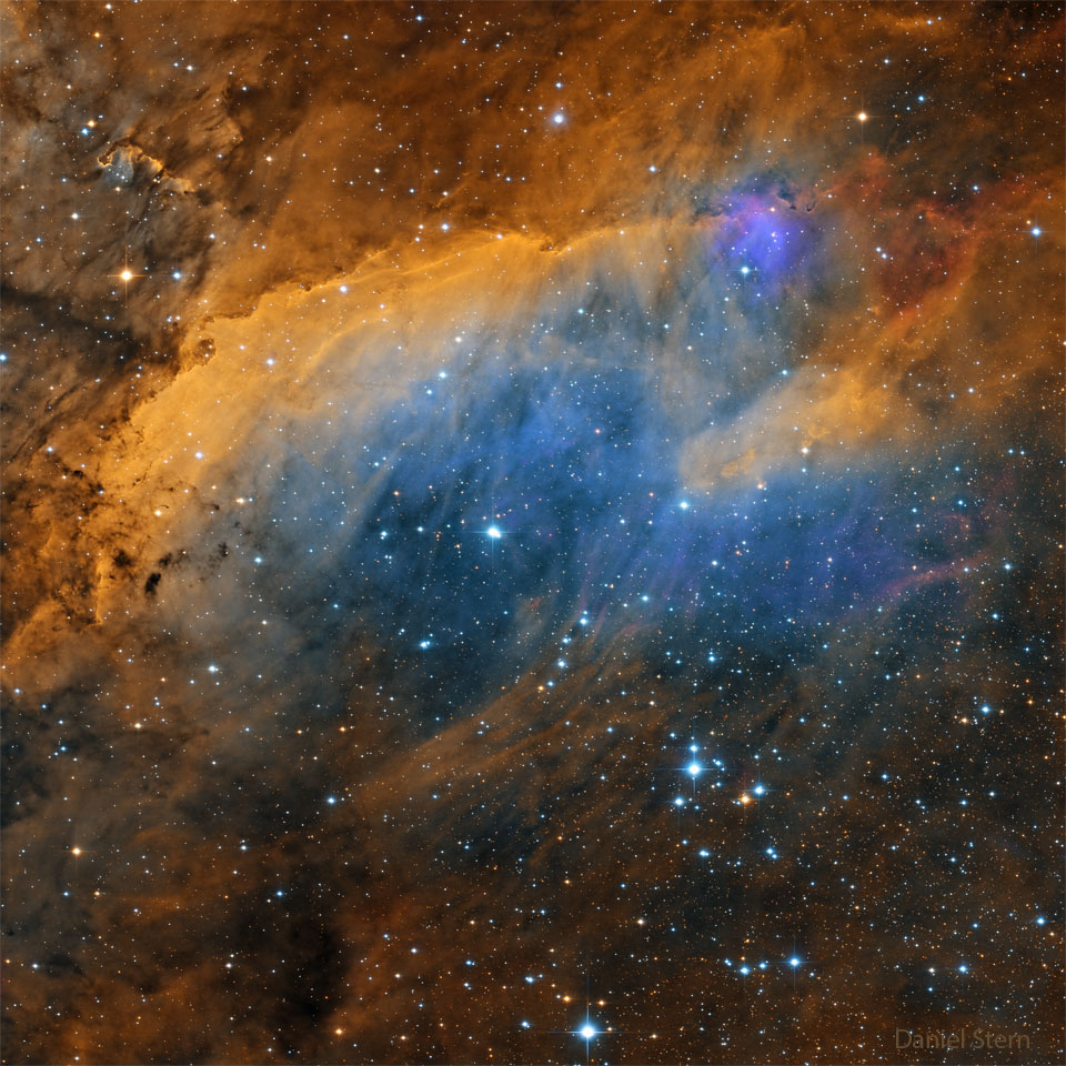 A sprawling nebula is pictured with gold tinted gas covering the   top, blue, the middle, and dark brown the bottom. Stars cover the frame  but are most prominent near the bottom.  Please see the explanation for more detailed information.