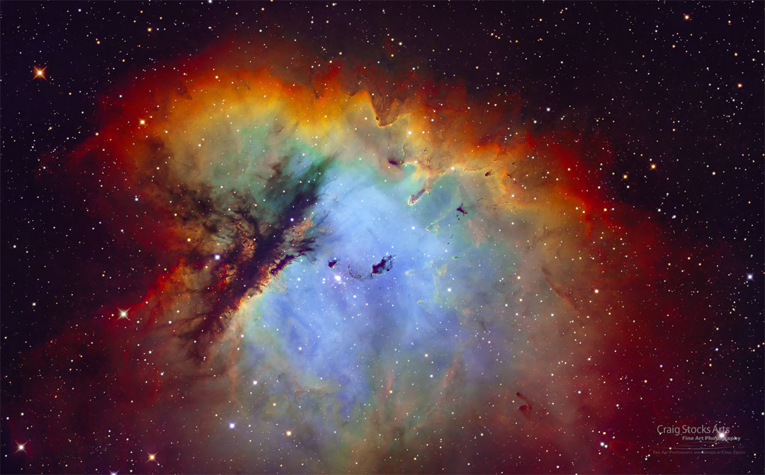 A nebula that appears blue in the middle and is surrounded by red-glowing gas is featured. Dramatic lanes of dark dust cut through the nebula's left side. A group of stars is visible toward the nebula's center. Please see the explanation for more detailed information.