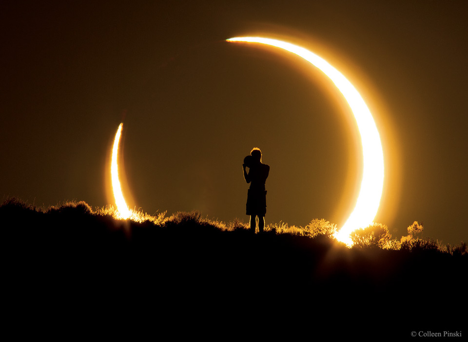 A person is seen standing at the top of a ridge. The person
appears as a silhouette onto the central dark region of an 
annular solar eclipse. The annular solar eclipse is a bright
ring with a large dark hole in the middle.
Please see the explanation for more detailed information.