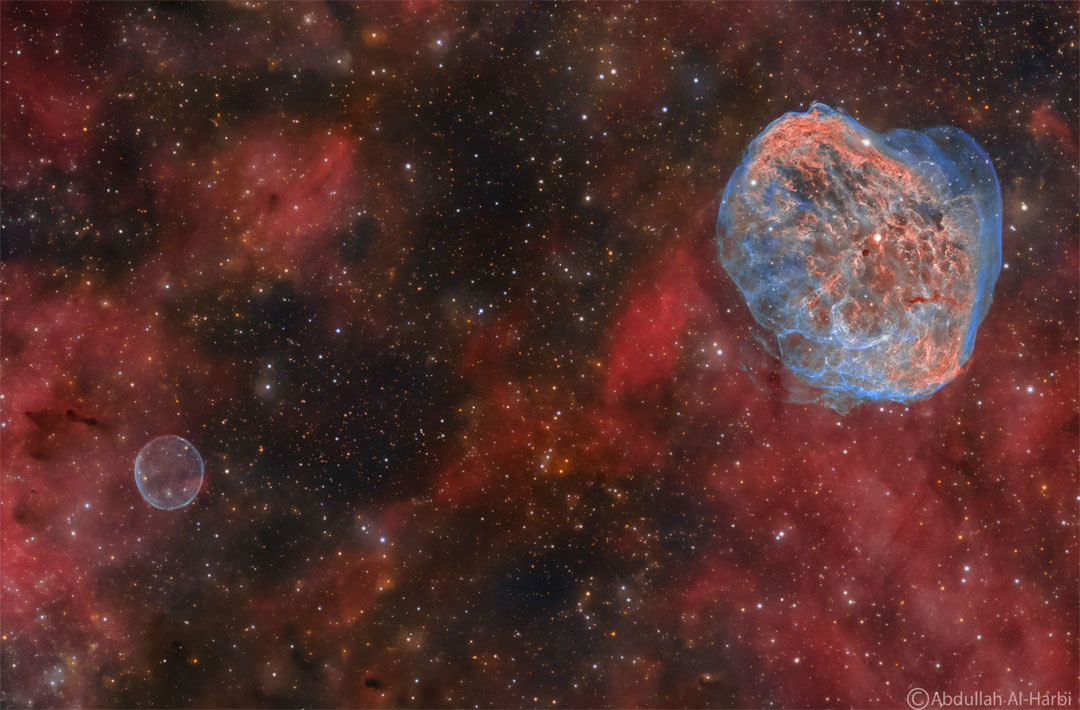 Red glowing gas is seen before a dark starfield. On the upper
right is a complicated filamentary nebula in blue and red. On the 
lower left is a simple circular nebula in blue.
Please see the explanation for more detailed information.