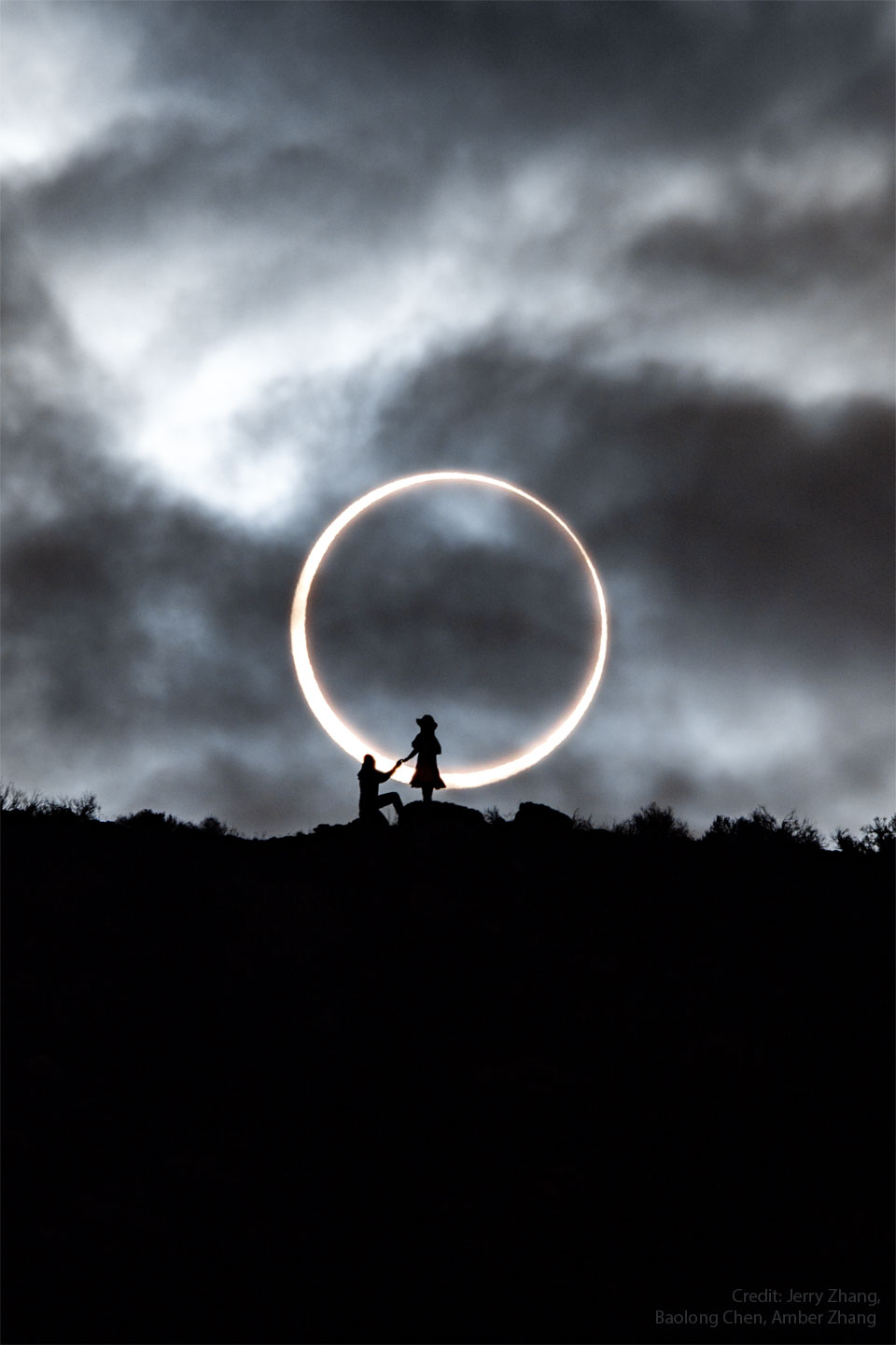 An annular solar eclipse appears in the background with the dark Moon appearing completely internal to the bright Sun. In the foreground is a ridge with the silhouettes of two people, one standing, and one kneeling. Please see the explanation for more detailed information.