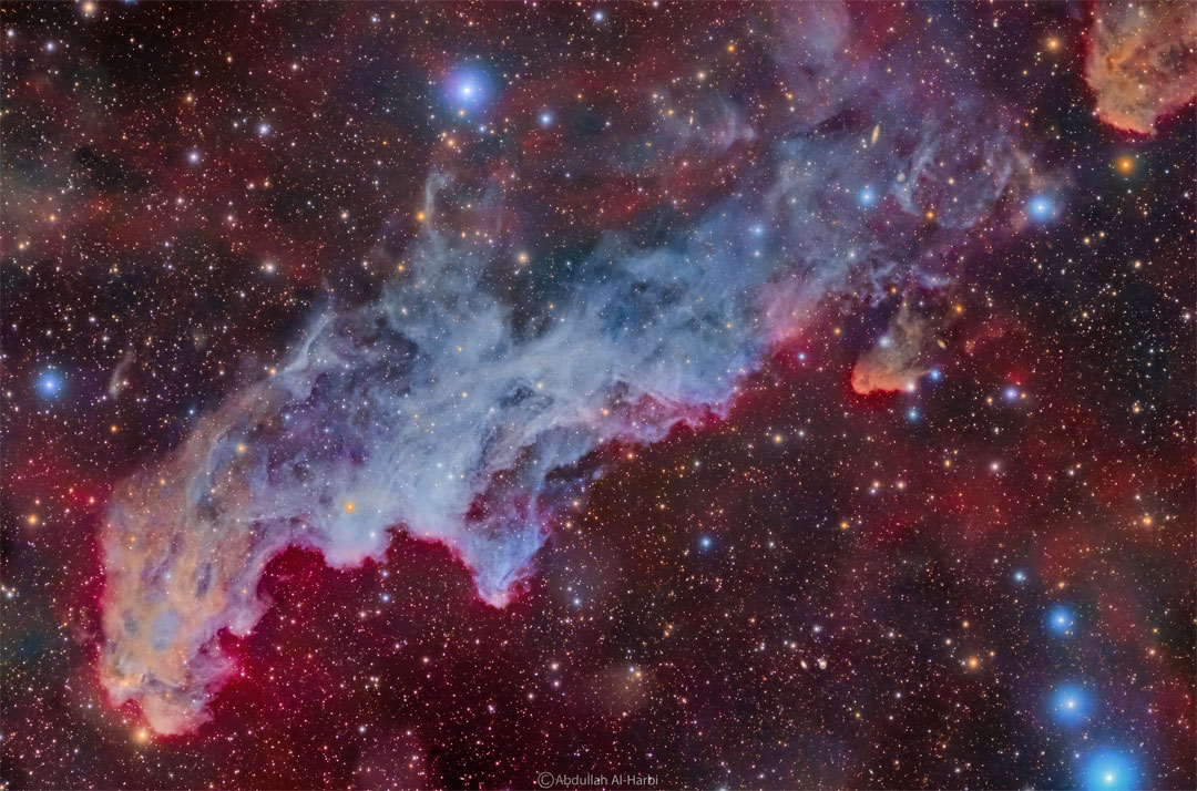 A colorful star field surrounds a big blue reflection nebula.  The nebula is elongated across the wide frame and said to   resemble the head of folklore-based witch.  Please see the explanation for more detailed information.