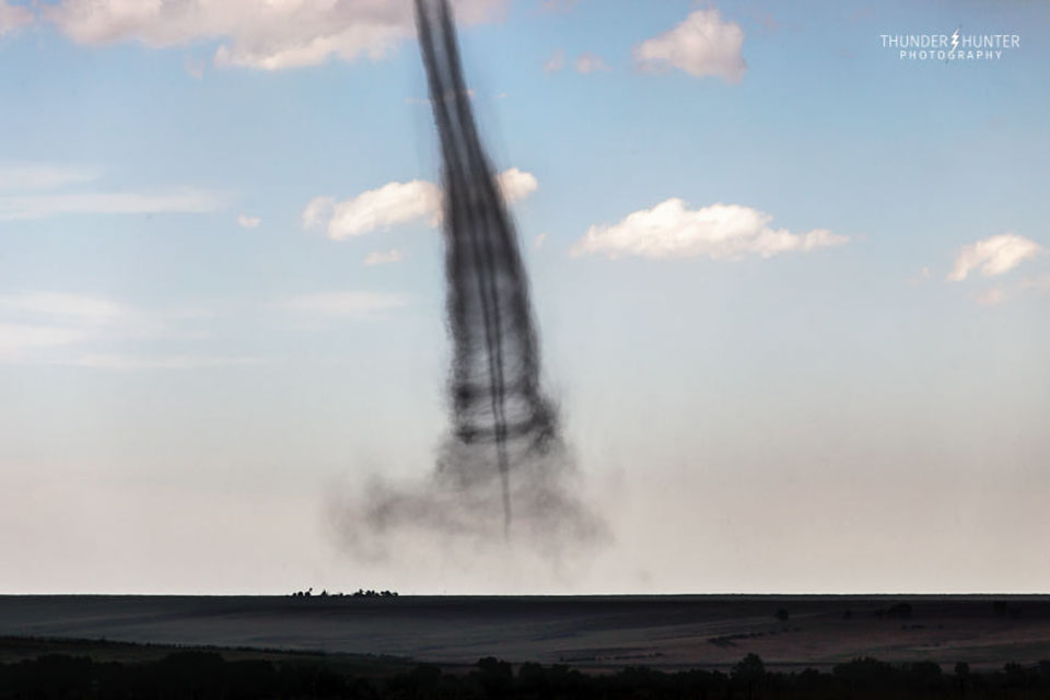 A funnel cloud is shown, but inside what appears to be 
a wider funnel cloud. A blue sky with a few white clouds is
seen in the background, while flat plains are seen in the 
foreground.
Please see the explanation for more detailed information.