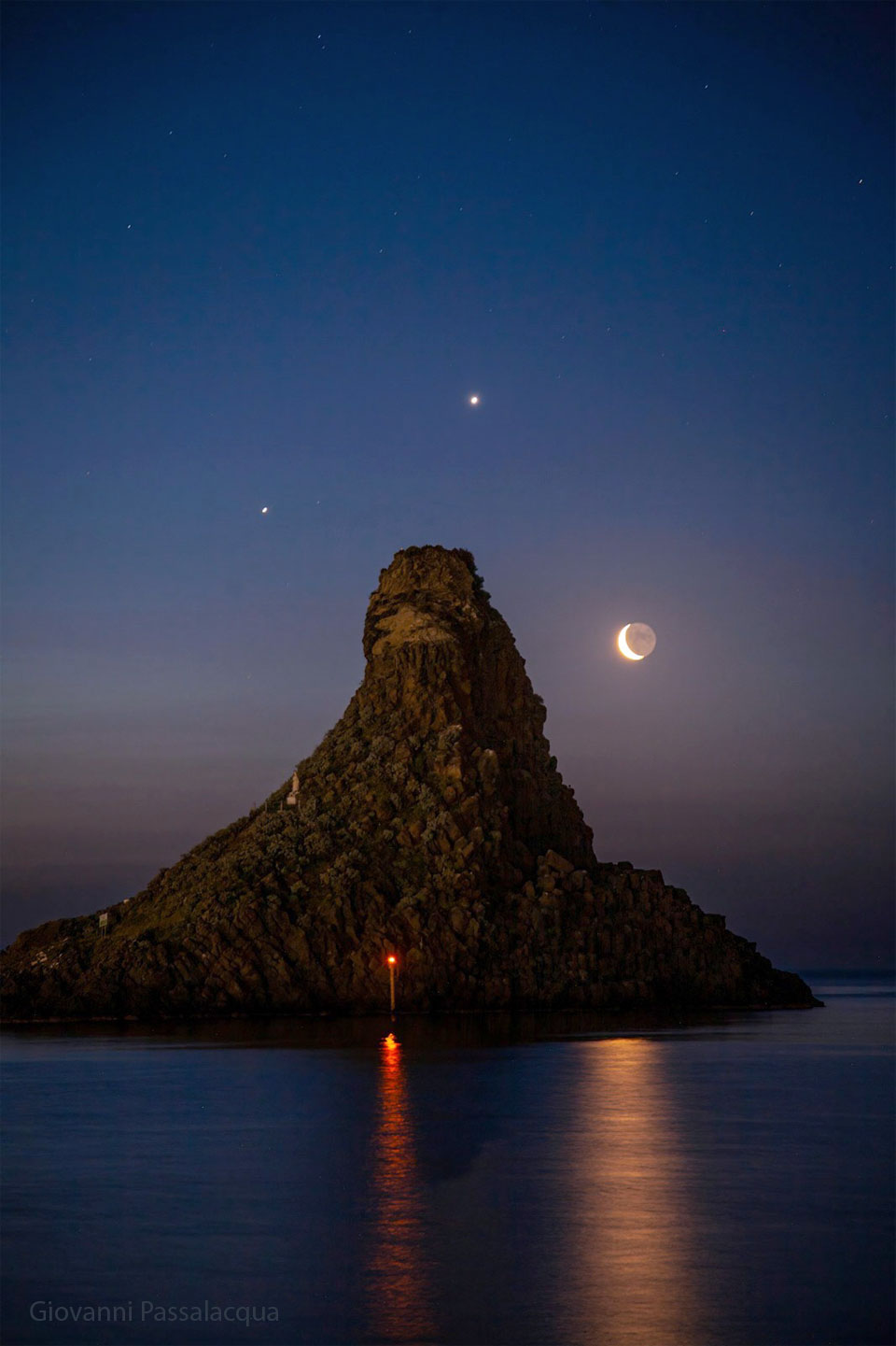 A seascape surrounds a large tree-covered hill. Surrounding the hill
in the night sky are three bright dots: the planets Jupiter, Venus,
and a crescent Moon.
Please see the explanation for more detailed information.