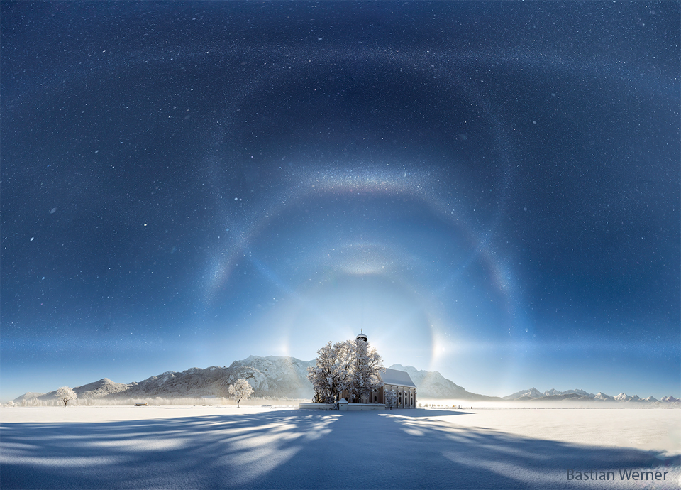 A building is seen from a distance on white snow and with
mountains in the background. An ice-crystal filled sky is seen above.
Superposed on the night sky are numerous curving whisps -- halos
of ice reflecting background moonlight.
Please see the explanation for more detailed information.