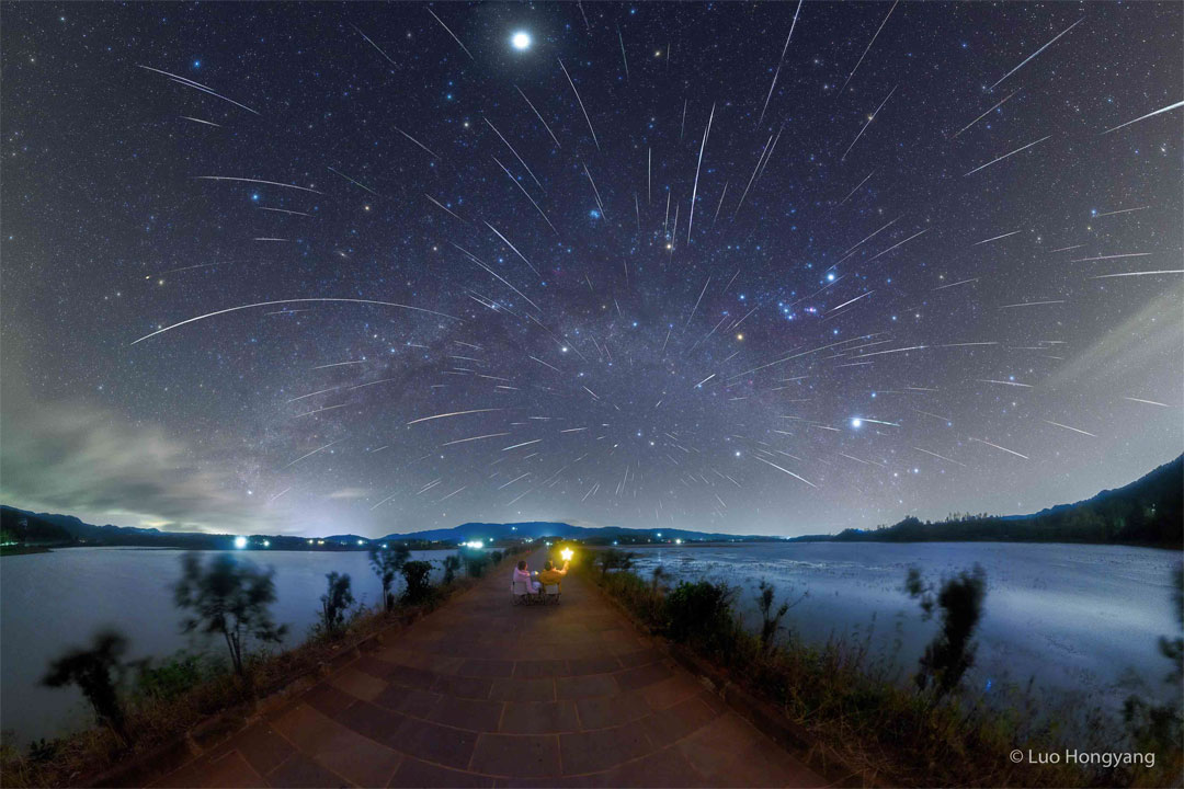 Two people are pictured from the back looking at a dark
star-filled sky. The sky is also filled with numerous streaks
caused by meteors from the Geminids meteor shower.
Please see the explanation for more detailed information.