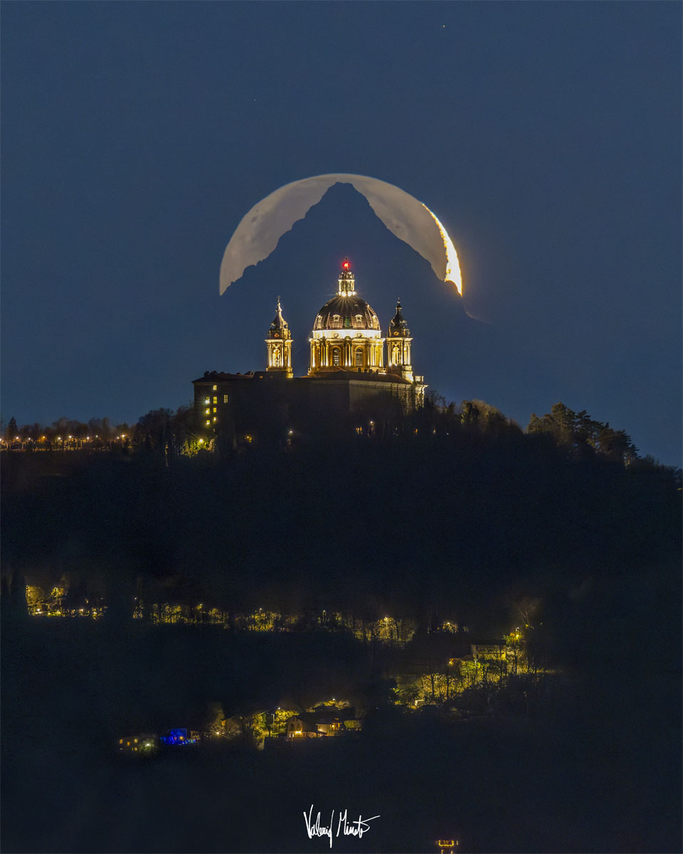 A tree-lined hill is shown topped by a majesticcathedral. Directly behind the cathedral is of a triangular-shaped mountain top. Directly behind the mountain is a crescent moon, although the exposure is long enough to see the rest of lunar circle. Please see the explanation for more detailed information.