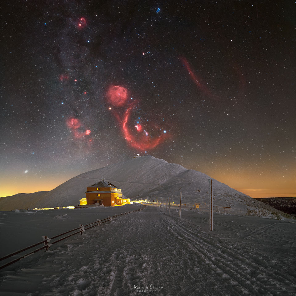 A snowy landscape is pictured with a big hill in the center.
Above the hill is a starfield with the stars and nebulae of the 
constellation Orion appearing, with the red glow of the 
nebulas in great contrast to the dark sky and bright snow.
Please see the explanation for more detailed information.