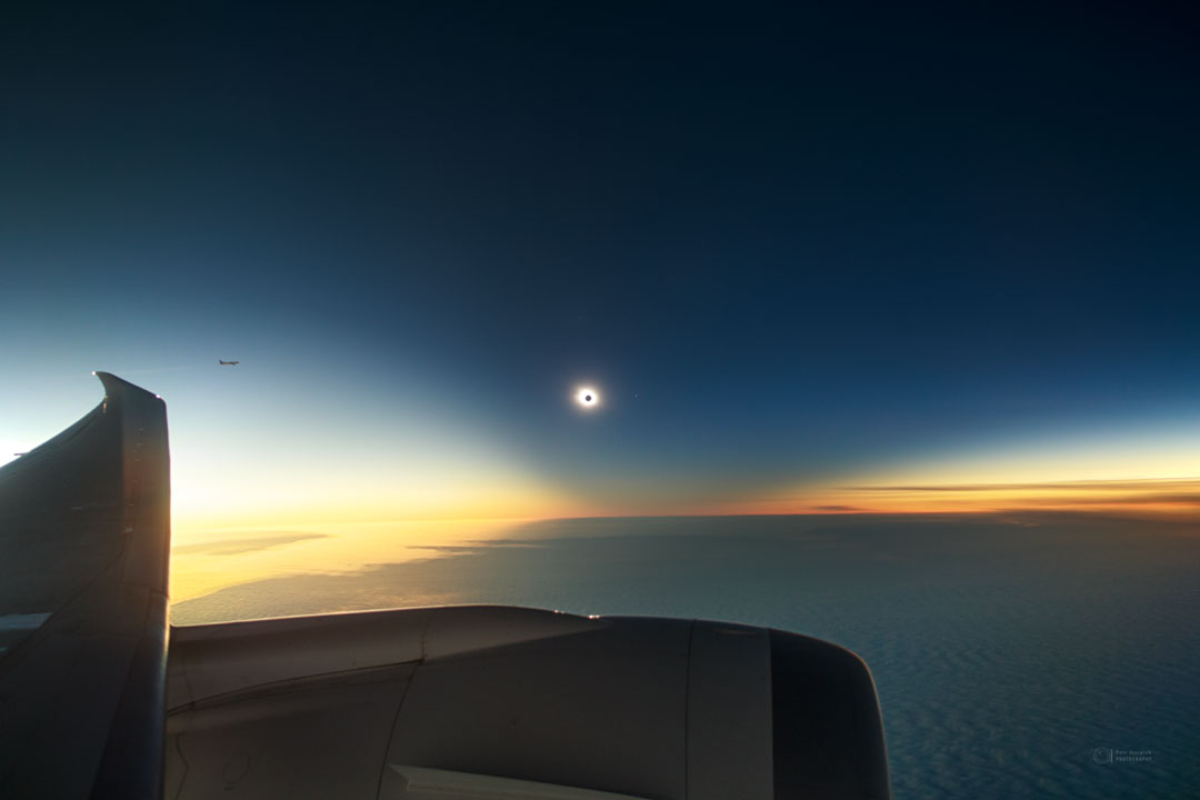 A totally eclipsed Sun is seen in the distance. Around
the eclipse is a dark region dipping down from above. Below
that are clouds and below that is the wing and engine of
an airplane.  
Please see the explanation for more detailed information.