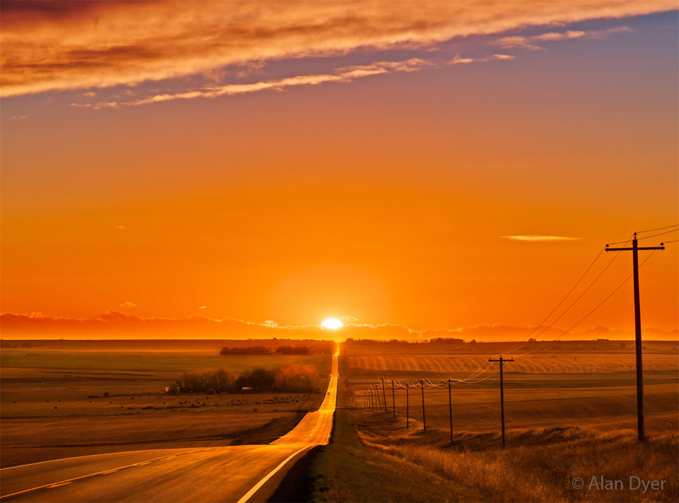 The sun sets in the distance at the horizon end of
a long road over open country. The sunset is very orange,
as is the surrounding sky. Telephone poles line the right
side of the road.
Please see the explanation for more detailed information.