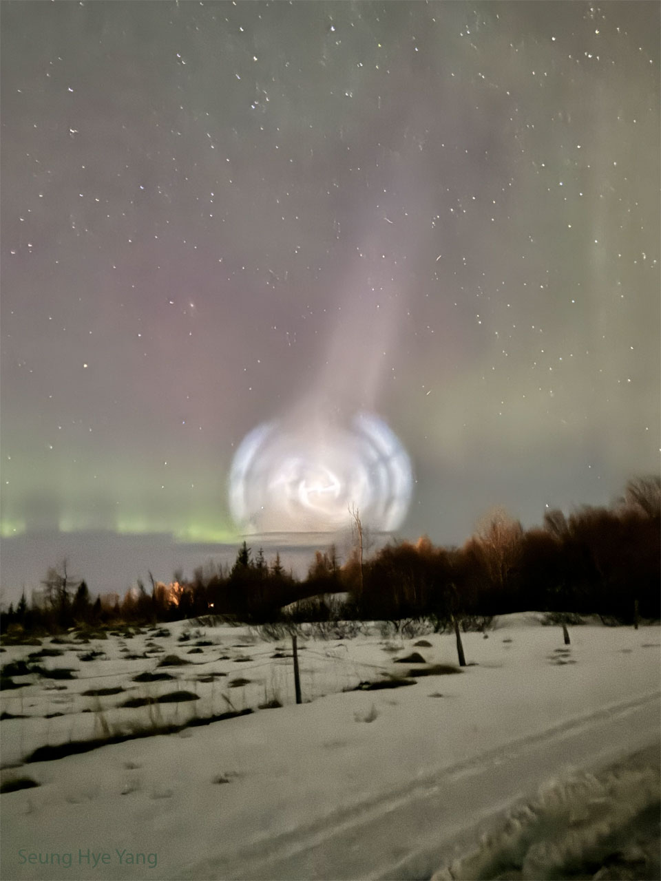 A field of snow is shown, lined with trees along the back.  Above the horizon is an unusual white spiral cloud.   Stars dot the background, and faint green and red aurora  are also visible.   Please see the explanation for more detailed information.