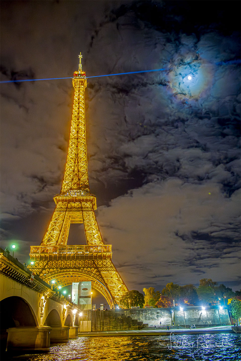 The famous Eiffel Tower in Paris, France is pictured 
on the left lit up in gold at night. A blue laser shines
out from the top. Clouds dot the background sky. The Moon
is also visible through the clouds, but is circled by 
colorful rings: a lunar corona.
Please see the explanation for more detailed information.