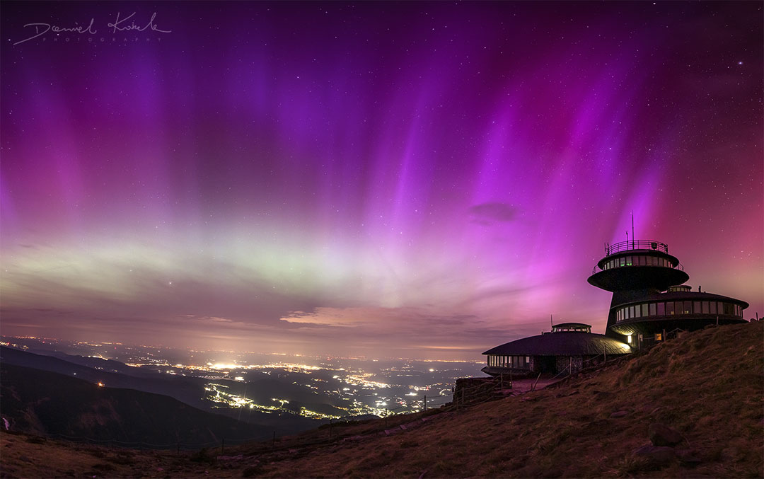 Purple striped aurora cover a star filled sky. Mountain
peaks are visible on the sides, as well as a futuristic
looking building on the right side. City lights are 
visible in the valley down below. 
Please see the explanation for more detailed information.
