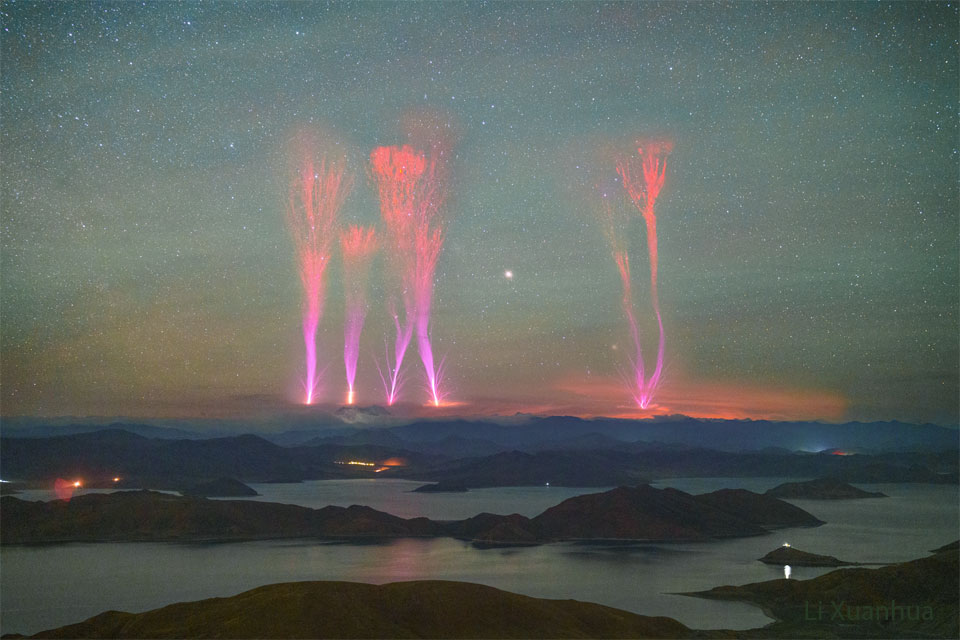 A landscape showing a night sky over distant mountains
is shown. Lakes dot the foreground in front of the mountains.
Extending from above the mountains into the night sky are 
six bright jets. The jets are violet at the bottom but red
at the top. 
Please see the explanation for more detailed information.