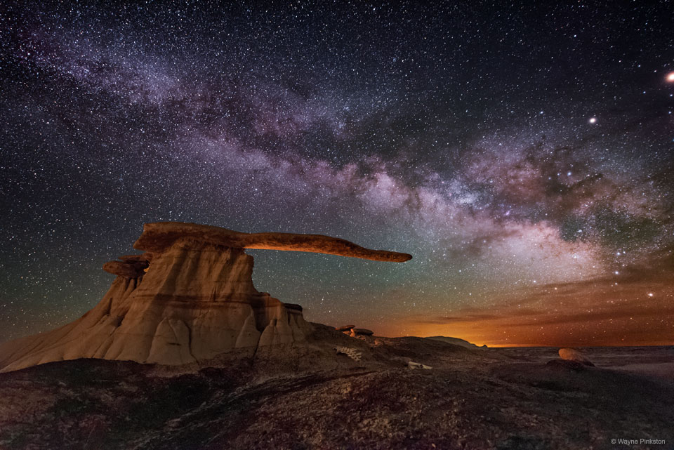 The band of the Milky Way runs across a night sky filled with stars. Colorful clouds are on the right horizon. A strange rock structure appears in the image center with a base and an extended arm that seems to point to the colorful horizon. Please see the explanation for more detailed information.