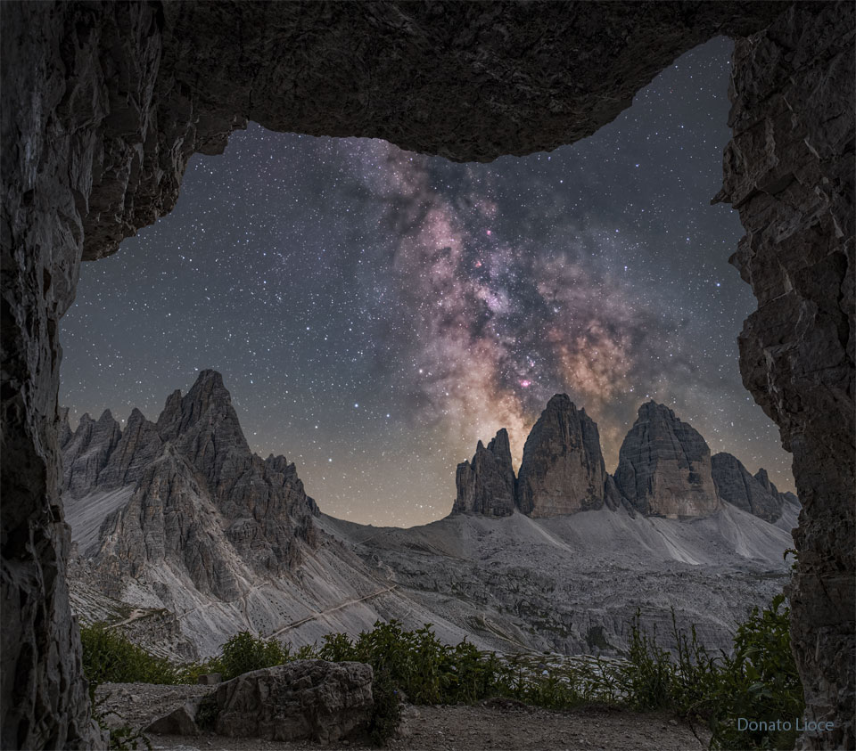 A starry sky is seen through a rectangular rock  opening. Three rocky peaks appear toward the right, while  other peaks appear toward the left. Rising above the   right peaks is the central band of our Milky Way Galaxy.  Please see the explanation for more detailed information.