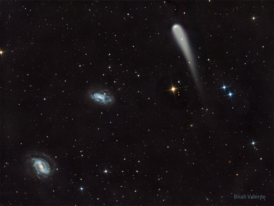 A picture shows a starfield with three prominent objects.  A blue spiral galaxy is on the lower left and another blue spiral  is just left of center. Toward the upper right is a light-colored  comet with a tail fading toward in the image bottom.  Please see the explanation for more detailed information.