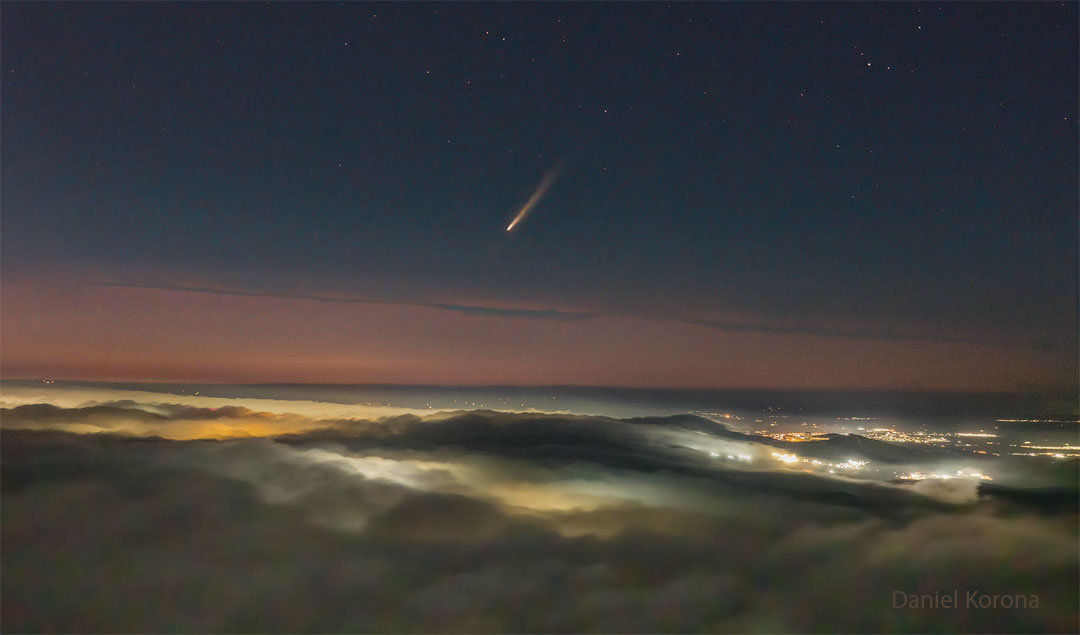 A picture shows clouds across the bottom and a dark night sky  across the top. In the middle is a band of orange sky. City lights  are visible on the right through gaps in the clouds. In the center  of the upper sky is a comet with its tail pointing toward the   upper right.   Please see the explanation for more detailed information.