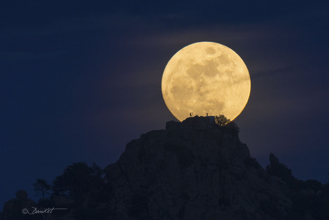 Earth's Moon is shown just beyond a rocky hill. The Moon is  near full phase. On the hill the silhouette of a person looking  through a telescope can be seen. A rollover darkens part of the   Moon that looks to some like a human face.  Please see the explanation for more detailed information.