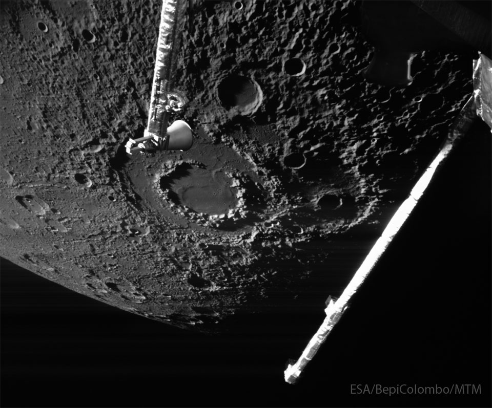 The cratered surface of a large body is shown: Mercury.  The largest feature visible is a large impact crater with two  rings, near the image center. Arms from the BepiColumbo   spacecraft that took the image are seen extending into the   image from the top and the right.   Please see the explanation for more detailed information.