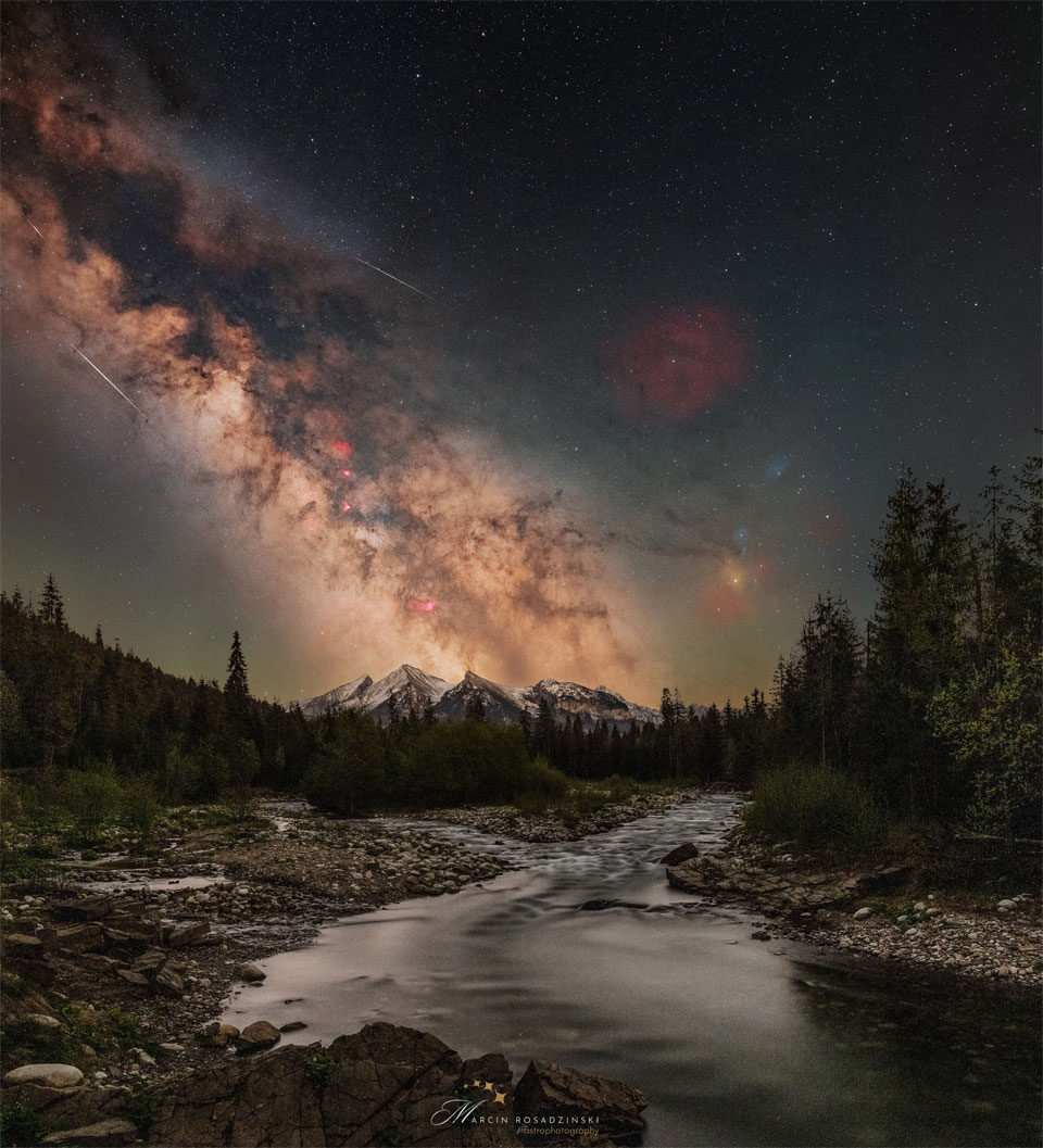 A starry sky is shown with the busy central band of   our Milky Way Galaxy showing diagonally from the upper left.  Mountains are on the horizon, with trees and a stream running  up from the foreground.   Please see the explanation for more detailed information.