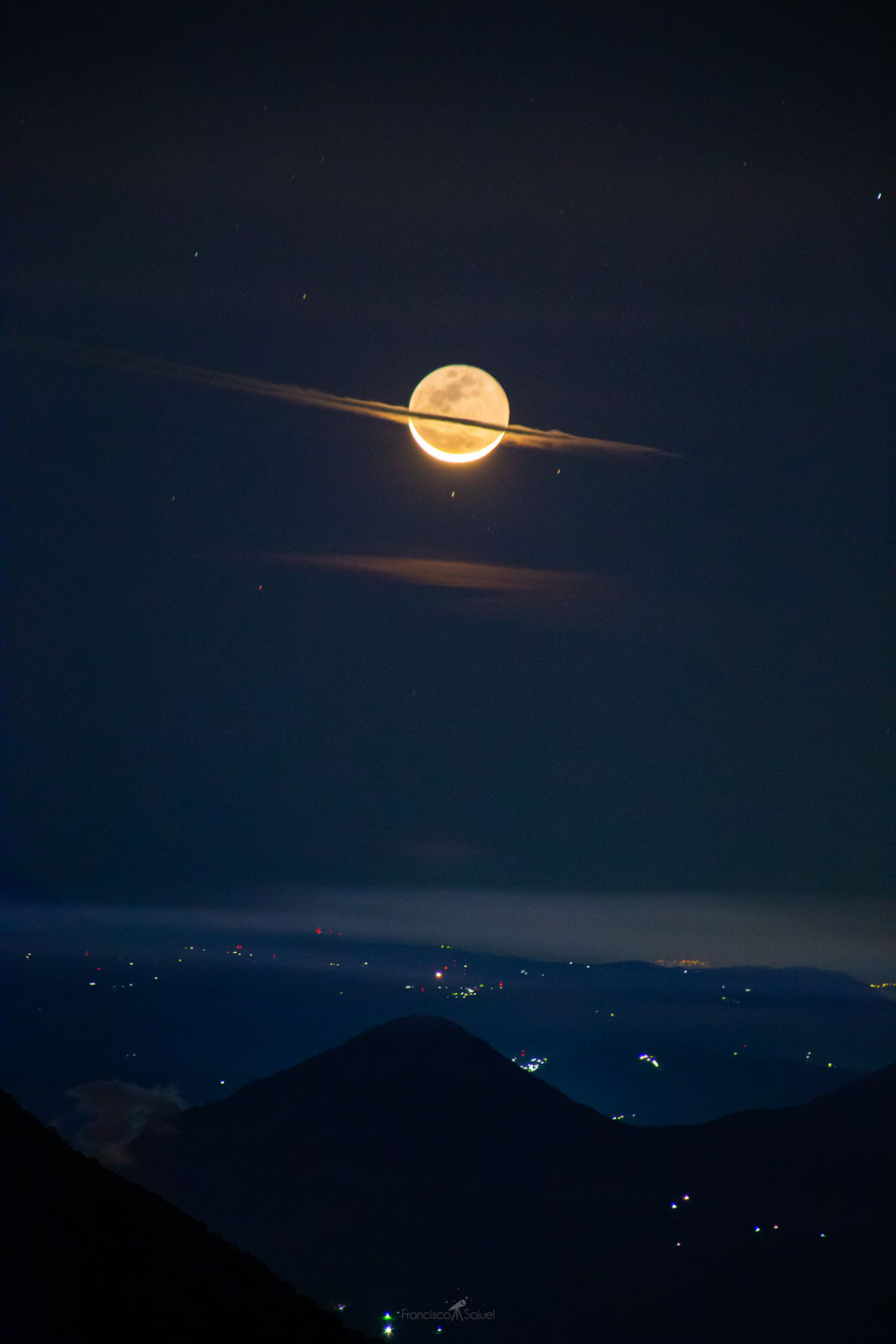 The featured image shows a crescent Moon over a city  and volcano with a flat cloud running through the center that  makes the Moon look a bit like the planet Saturn.  Please see the explanation for more detailed information.