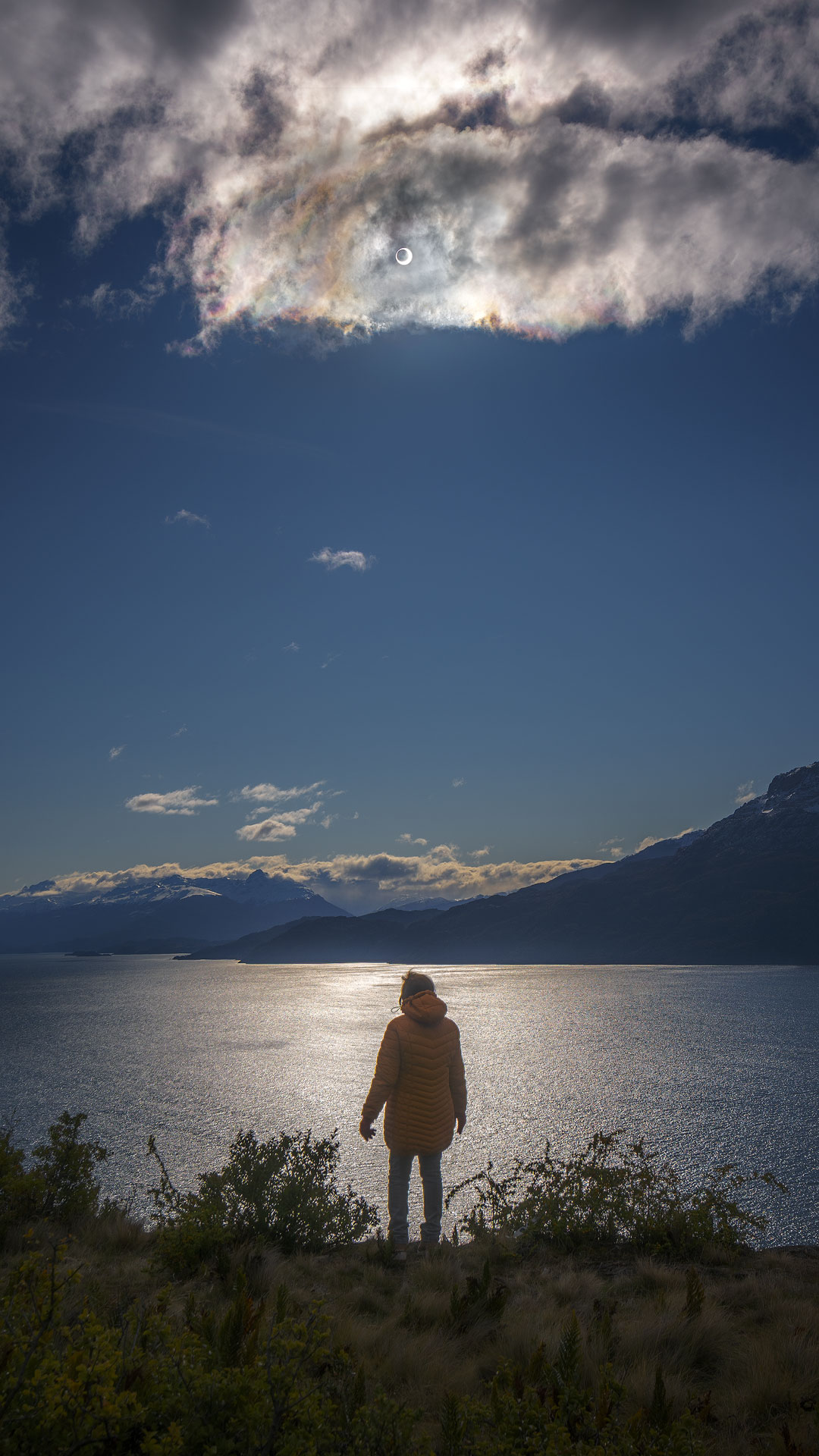 A person stands overlooking a lake. The sun is high in a partly cloudy sky. If you look closely at the Sun, you'll see a dark spot in the center. This is the moon during an annular solar eclipse. See description for details.