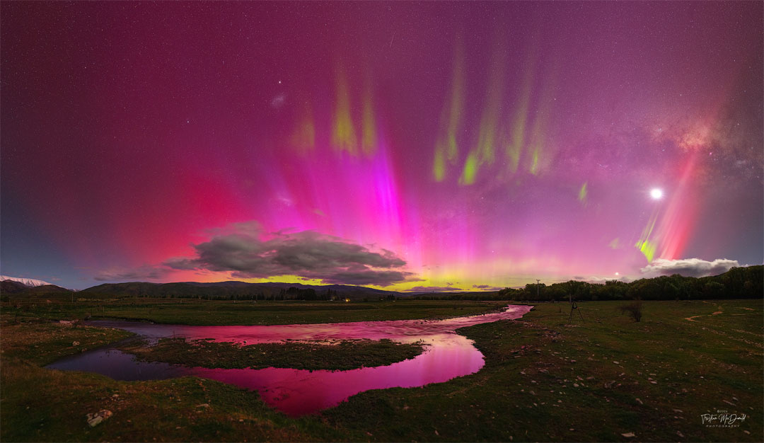 A night sky is shown that appears mostly red due to pervasive  aurora. In the foreground is covered by watery grasslands. Clouds   are visible above the horizon. Thin green aurora are visible   toward the top of the frame. In the background one can find the Moon,  the LMC, SMC, Venus, a meteor, and the band of our Milky Way galaxy.  Please see the explanation for more detailed information.