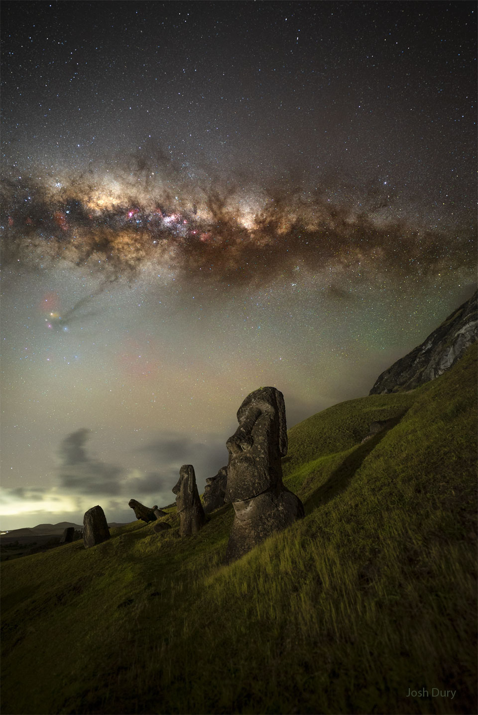 A grassy hill appears in the foreground with tall statues   of human heads embeddd. High overhead the central band of the   Milky Way galaxy crosses horizontally. Above the Milky Way is  a dark sky filled with stars.   Please see the explanation for more detailed information.